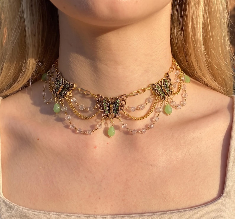 May include: A gold-toned choker necklace featuring three butterfly charms with green accents. The design includes delicate gold chains draped between the butterflies, interspersed with clear beads and small, dangling light green teardrop-shaped stones.