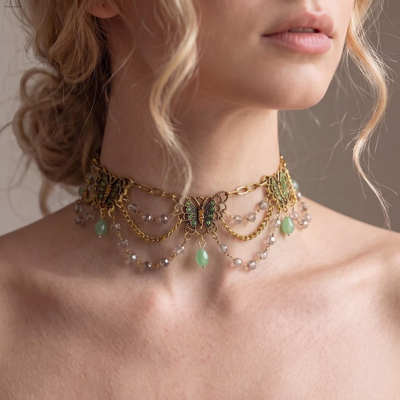 May include: A gold-toned choker necklace featuring three intricate butterfly charms with green accents. The design includes delicate gold chains draped between the butterflies, interspersed with small, translucent grey beads and dangling light green teardrop-shaped stones.
