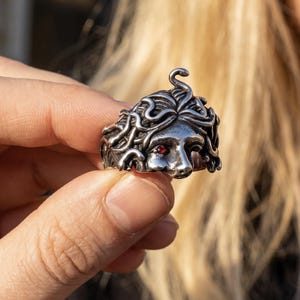 Könnte beinhalten: Ein silberfarbener Metallring mit einem skulptierten Gesicht, detailliertem schlangenartigem Haar und einem einzelnen kleinen, runden, roten Edelstein in einer Augenhöhle. Das Design weist ein dunkles, oxidiertes Finish in den Vertiefungen auf.