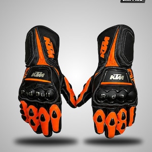 May include: A pair of black and orange motorcycle gloves featuring the KTM logo in orange on the wrist and white on the knuckle guard. The gloves include black knuckle protection with the text Red Bull printed in silver on the left glove.