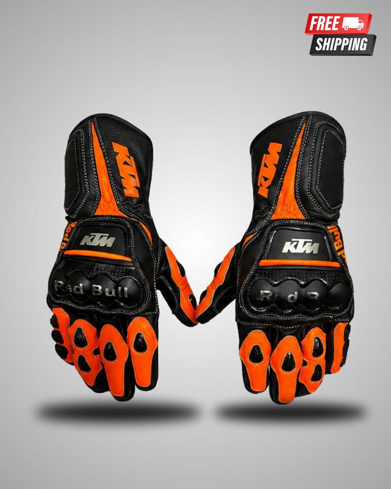 May include: A pair of black and orange motorcycle gloves featuring the KTM logo in orange on the wrist and white on the knuckle guard. The gloves include black knuckle protection with the text Red Bull printed in silver on the left glove.