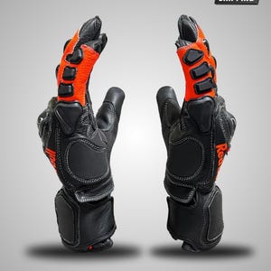 May include: A pair of black and orange leather motorcycle gloves featuring protective hard-shell knuckle guards, reinforced palm padding, and white contrast stitching. The gloves display the text Red Bull in orange on the side of the wrist.