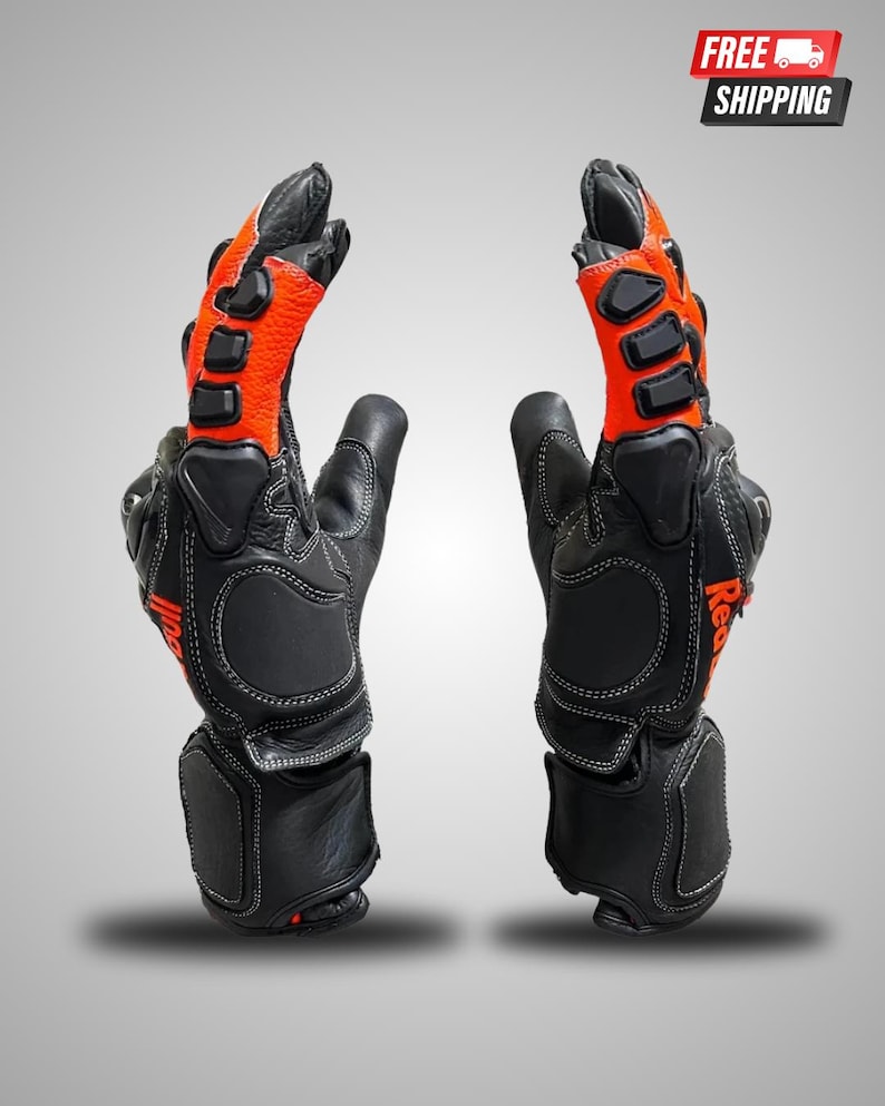 May include: A pair of black and orange leather motorcycle gloves featuring protective hard-shell knuckle guards, reinforced palm padding, and white contrast stitching. The gloves display the text Red Bull in orange on the side of the wrist.