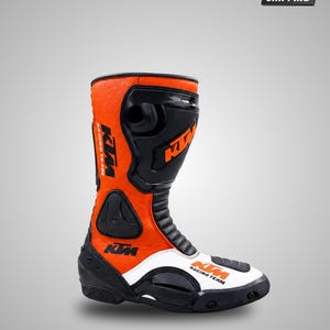 May include: A high-top motorcycle boot in orange, black, and white. The boot features the KTM Racing Team logo in black and orange on the side and toe. It includes molded black protective panels on the shin, ankle, and heel, with a textured finish.