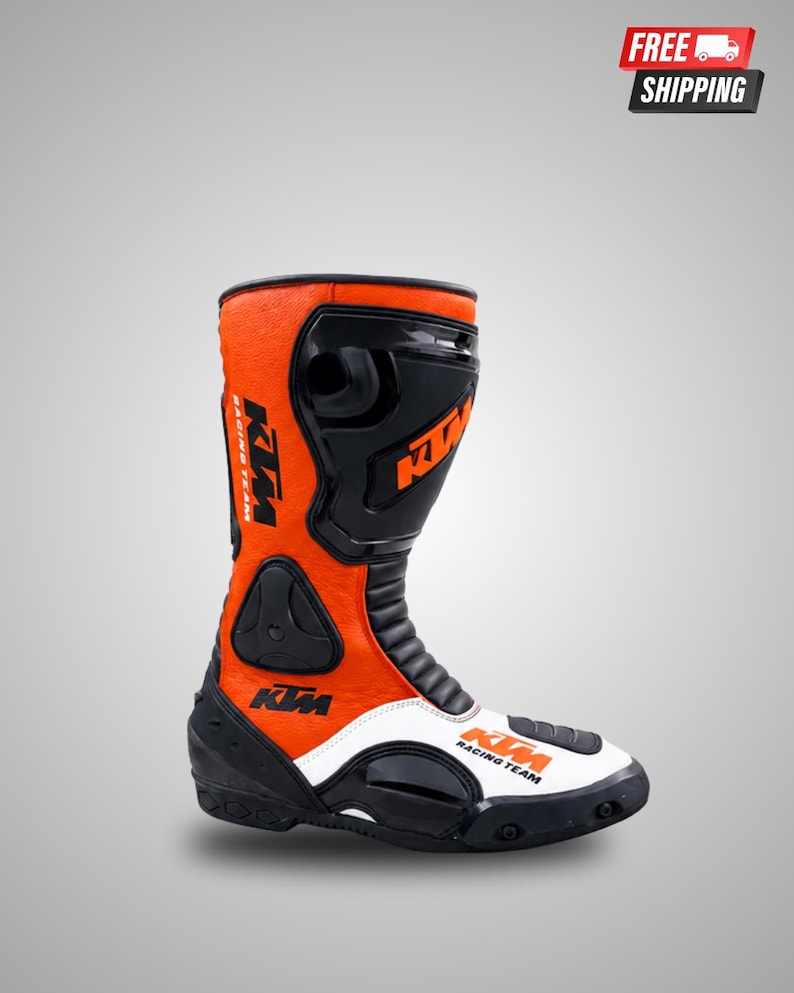 May include: A high-top motorcycle boot in orange, black, and white. The boot features the KTM Racing Team logo in black and orange on the side and toe. It includes molded black protective panels on the shin, ankle, and heel, with a textured finish.