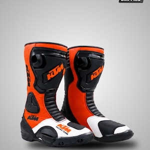 May include: A pair of motorcycle boots in bright orange, black, and white. The boots feature the KTM logo in orange on the black shin guard and the text KTM RACING TEAM on the white toe section. They include black accordion panels and protective gear.