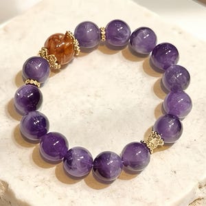 May include: A beaded bracelet featuring large, round, polished amethyst stones in varying shades of purple. The bracelet includes a central, ornate, gold-colored bead and gold-colored accents between the amethyst beads. The bracelet is displayed on a light-colored surface.