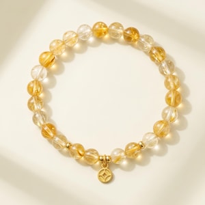 Pode incluir: Uma pulseira de contas com contas de quartzo amarelo dourado e transparente. A pulseira tem um pingente dourado com um design geométrico. As contas são enfiadas com detalhes dourados. A pulseira é exibida em uma superfície branca.