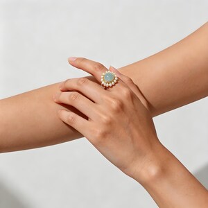 May include: A gold ring featuring a central round light blue cabochon stone surrounded by a halo of small white pearls. The ring is worn on a finger, showcasing the circular arrangement of the pearls around the smooth, pale blue gemstone.