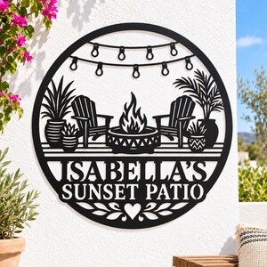 May include: A circular black metal wall sign featuring a cutout design of two chairs, a fire pit, potted plants, and string lights. The text ISABELLA'S SUNSET PATIO is centered below the scene, with a heart and leaf motif at the bottom.
