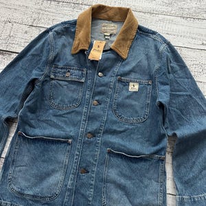 May include: A blue denim chore jacket featuring a tan corduroy collar, long sleeves, and a button-front closure. The jacket includes two chest pockets and two large lower pockets, with a Polo Ralph Lauren brand tag attached to the collar.