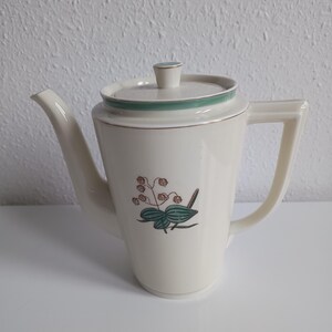 Puede incluir: Tetera de cerámica color crema con tapa y asa. La tetera presenta una banda verde turquesa alrededor de la tapa y un diseño floral en verde turquesa, marrón y verde. El pico es curvo y el asa rectangular.