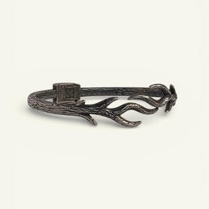May include: A silver-toned bracelet with a textured, branch-like design. The bracelet features a clasp with a square design and ends with a stylized antler motif. The overall aesthetic is rustic and nature-inspired.