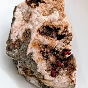May include: A rough-hewn mineral specimen with a mix of colors. The rock features areas of light pink, brown, and gray, with small clusters of deep red crystals. The overall texture is coarse and uneven, with visible crystalline structures.