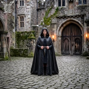 May include: A person wearing a black hooded cloak with a belt, standing on a cobblestone path in front of a castle. The cloak is long and flowing, and the person is facing the camera. The background includes stone walls, arched doorways, and torches.