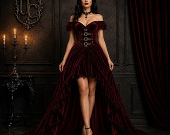May include: A deep burgundy velvet high-low dress featuring an off-the-shoulder neckline, ruffled sleeves, and a bodice adorned with four vertical buckled straps. The skirt has a shorter front hemline and a long, flowing train with ruffled trim.