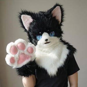 以下が含まれることがあります： 大きな青い目とピンクの内側の耳を持つ、白と黒の猫のファースーツヘッド。 ファースーツはピンクの肉球パッドで手を振っています。 猫の毛皮はふわふわで詳細で、リアルな外観を作り出しています。