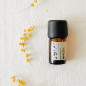 May include: A small amber glass essential oil bottle with a black cap. The label reads "DREAMY MOON" and lists the top and middle notes. Yellow flowers are scattered around the bottle on a white textured surface.