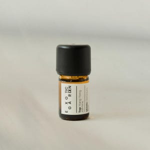 May include: A small amber glass bottle with a black cap. The label reads "EXOTIC GARDEN" and lists the top and middle notes of the fragrance. The bottle contains a liquid, likely an essential oil or perfume.
