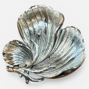 May include: A silver-colored, leaf-shaped decorative bowl. The bowl has a textured surface with detailed veining, and a small branch-like accent. The bowl is likely made of metal and is designed to hold small items or serve as a decorative piece.