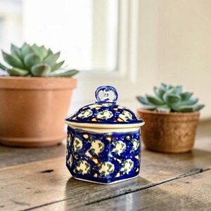 May include: A small, hexagonal ceramic jar with a matching lid, featuring a vibrant blue, white, and brown patterned design. The jar has a decorative loop handle on top of the lid and a smooth, glossy finish, showcasing intricate hand-painted details.