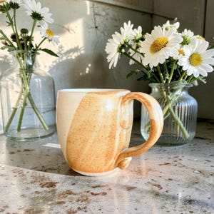 May include: A handmade ceramic mug with a tan and cream color scheme. The mug has a rounded handle and a unique design. The mug is sitting on a stone surface, with vases of daisies in the background.
