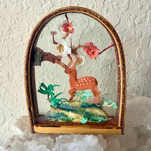 May include: An arched wooden display case containing a three-dimensional diorama. Inside, a brown spotted deer stands on a textured green and yellow base beneath a branch with white and pink blossoms and green foliage accents.