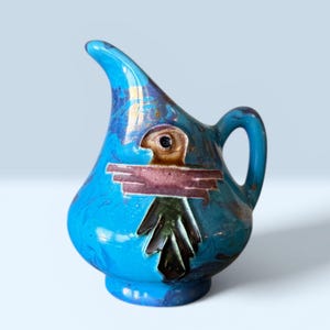 May include: A turquoise ceramic pitcher with a bird design. The pitcher has a curved spout and a handle. The bird design is brown, pink, and green. The pitcher has gold accents and a marbled effect.