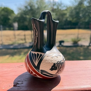 May include: A black, red, and cream-colored ceramic vessel featuring a double-spout design connected by a bridge. The surface is decorated with etched geometric patterns, a feather motif on the neck, and a bear figure on a textured cream panel.