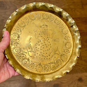 May include: A round brass tray with a scalloped edge featuring an engraved central figure of a deity holding a staff and a small bowl, surrounded by a circular border of stylized floral patterns etched into the metallic gold surface.