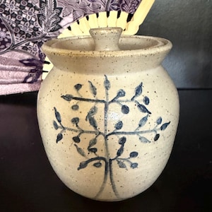 May include: A speckled beige ceramic jar with a matching lid and a hand-painted blue tree motif on the front. The jar features a rounded body, a wide rim, and a small knob handle on the lid. The surface has a textured, earthy appearance.
