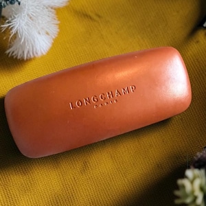 May include: A rectangular, burnt orange hard-shell glasses case with rounded corners. The brand name LONGCHAMP PARIS is embossed in the center of the top surface in a clean, sans-serif font. The case has a smooth, matte finish throughout.