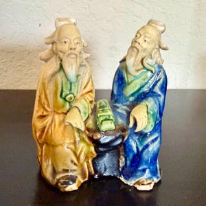 May include: A ceramic figurine depicting two elderly men in traditional robes. One figure wears yellow and green robes, the other blue and green. They are seated, with a small object between them. The figurine has a glazed finish.