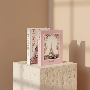 May include: Two decorative books with the word "PARIS" printed on the spines. One book is white with gold lettering, and the other is pink with an image of the Eiffel Tower. The books are displayed on a textured, square pedestal.