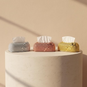 May include: Three decorative tissue box covers in silver, rose gold, and gold. Each box features a floral design and is filled with white tissues. The boxes are displayed on a white, rounded surface against a neutral background.