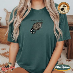 May include: Teal-colored t-shirt featuring a cartoon opossum holding a knife. The opossum has gray fur, a striped tail, and is positioned below the text "i require snacks". The shirt has a relaxed fit and short sleeves.