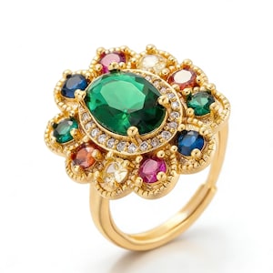 Puede incluir: Anillo dorado con una gran gema verde esmeralda ovalada en el centro, rodeada de gemas más pequeñas y coloridas. El anillo tiene una banda ajustable y un diseño floral. El diseño del anillo está inspirado en flores.