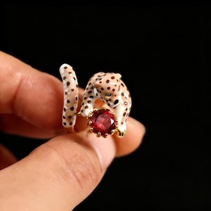 Könnte beinhalten: Goldener Ring mit einem Leoparden-Design, schwarzen Flecken und einem roten Edelstein. Der Leopard scheint den Edelstein zu halten. Der Schwanz des Rings umschlingt den Finger. Der Ring vor schwarzem Hintergrund.
