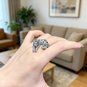 May include: A silver-toned ring in the shape of a leopard, encrusted with clear and black gemstones. The leopard is depicted in a crouching pose, with its head and paws visible. The ring is worn on a finger.