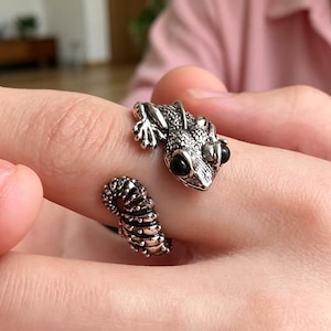 May include: A silver-colored ring in the shape of a gecko, with black eyes. The ring wraps around the finger, with the gecko's head and tail on opposite sides. The ring is detailed with scales and textured patterns.