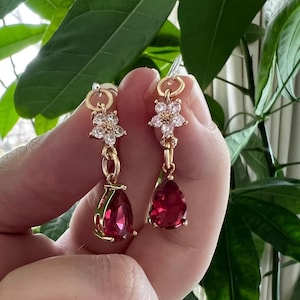 May include: A pair of gold-toned drop earrings. Each earring features a flower-shaped top with clear crystals, a chain, and a teardrop-shaped, deep red gemstone. The earrings are held up against a background of green leaves.