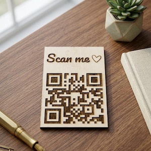 Könnte beinhalten: Eine Holzkarte mit einem QR-Code und dem Text "Scan me" und einem Herzsymbol. Die Karte liegt auf einer Holzoberfläche, neben einem goldenen Stift, Büroklammern, einer kleinen Sukkulentenpflanze in einem geometrischen Topf und einem Buch.