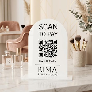 Op de afbeelding: Een wit boogvormig acrylbord met zwarte tekst die SCAN TO PAY leest, een QR-code, de tekst Pay with PayPal en de bedrijfsnaam RIMA BEAUTY STUDIO. Het bord is gemonteerd op een rechthoekige voet en toont een duidelijke en strakke typografie.