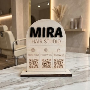 Puede incluir: Un letrero de madera arqueado con acabado en madera clara y base oscura. El letrero presenta el texto negro MIRA HAIR STUDIO, tres iconos que representan un calendario, una cámara y una estrella, y tres códigos QR etiquetados BOOK NOW, FOLLOW US, REVIEW US.