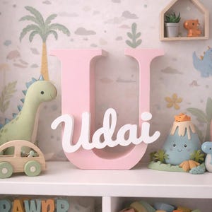 Könnte beinhalten: Ein rosa Holzbuchstabe "U" mit dem Namen "Udai" in weißer Schrift, ausgestellt auf einem weißen Regal. Das Regal ist mit Spielzeug zum Thema Dinosaurier dekoriert, darunter ein Plüschdinosaurier, ein Holzauto und ein Vulkan-Spielzeug.