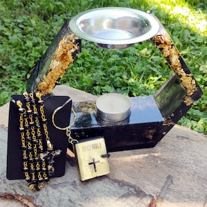 Può includere: Un oggetto decorativo nero e oro con una ciotola d'argento, una piccola candela, un rosario, un portachiavi con una croce e una Bibbia in miniatura. La Bibbia ha le parole "HOLY BIBLE" e una croce sulla copertina.