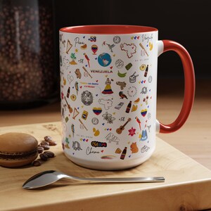 Könnte beinhalten: Eine weiße Keramiktasse mit rotem Innenraum und Henkel, verziert mit farbenfrohen Illustrationen zum Thema Venezuela. Das Design umfasst das Wort "Venezuela", Karten und verschiedene Symbole. Ein Löffel und ein Macaron befinden sich auf einer Holzoberfläche.