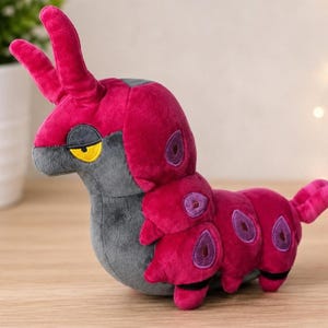 May include: A plush toy shaped like a segmented creature with a dark grey face, yellow eyes, and two long antennae. The body is vibrant magenta with purple oval markings along the sides and a small tail. The texture is soft and velvety throughout.