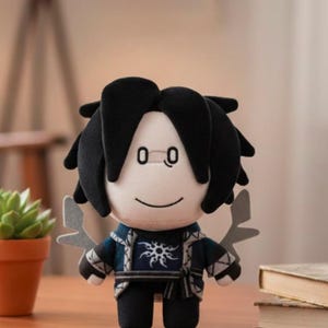 May include: A plush doll with black hair and a smiling face. The doll wears a dark blue jacket with a white sunburst design and black pants. Decorative gray wings are attached to the doll's back. The doll is standing in front of a blurred background.