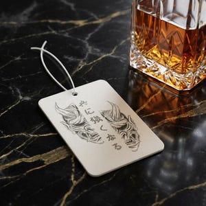 May include: A rectangular white air freshener featuring a black line art illustration of two Hannya masks facing each other with Japanese text in the centre. A thin white elastic string is attached to the top for hanging purposes.